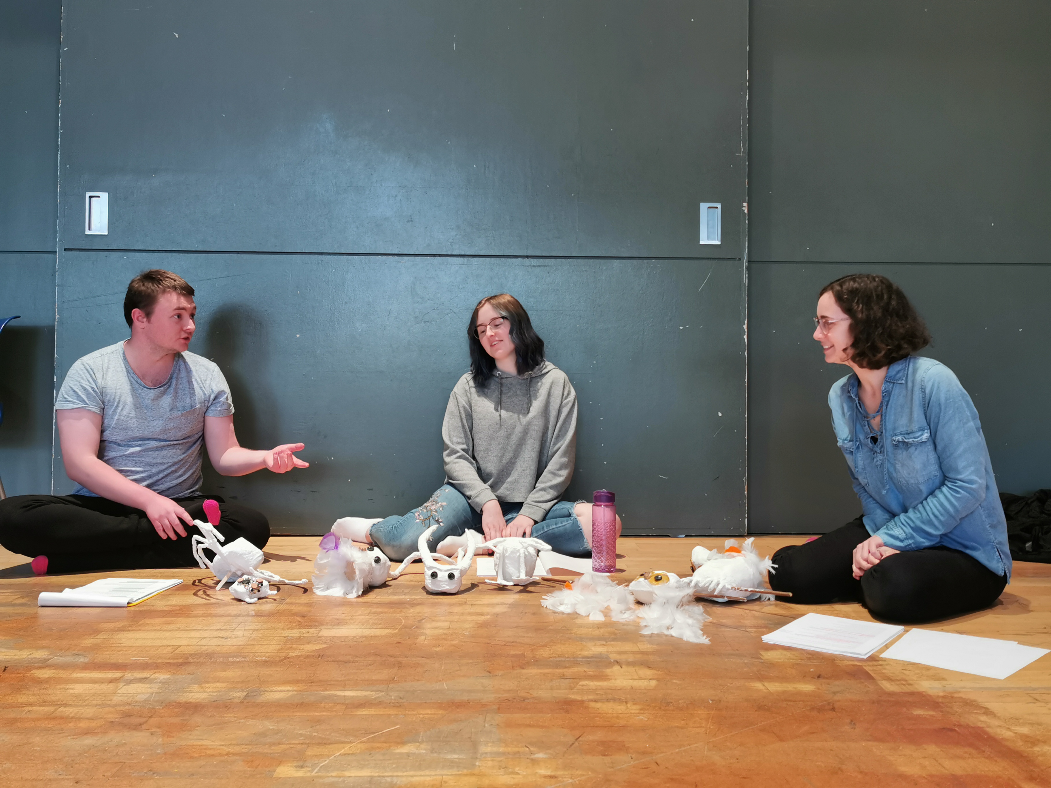 Cast members: John Tulloch, Heather MacDonald and Alina Ben Larbi sit in a semi-circle with scripts and puppet parts surrounding them. John narrates to Heather and Alina who listen, smiling.