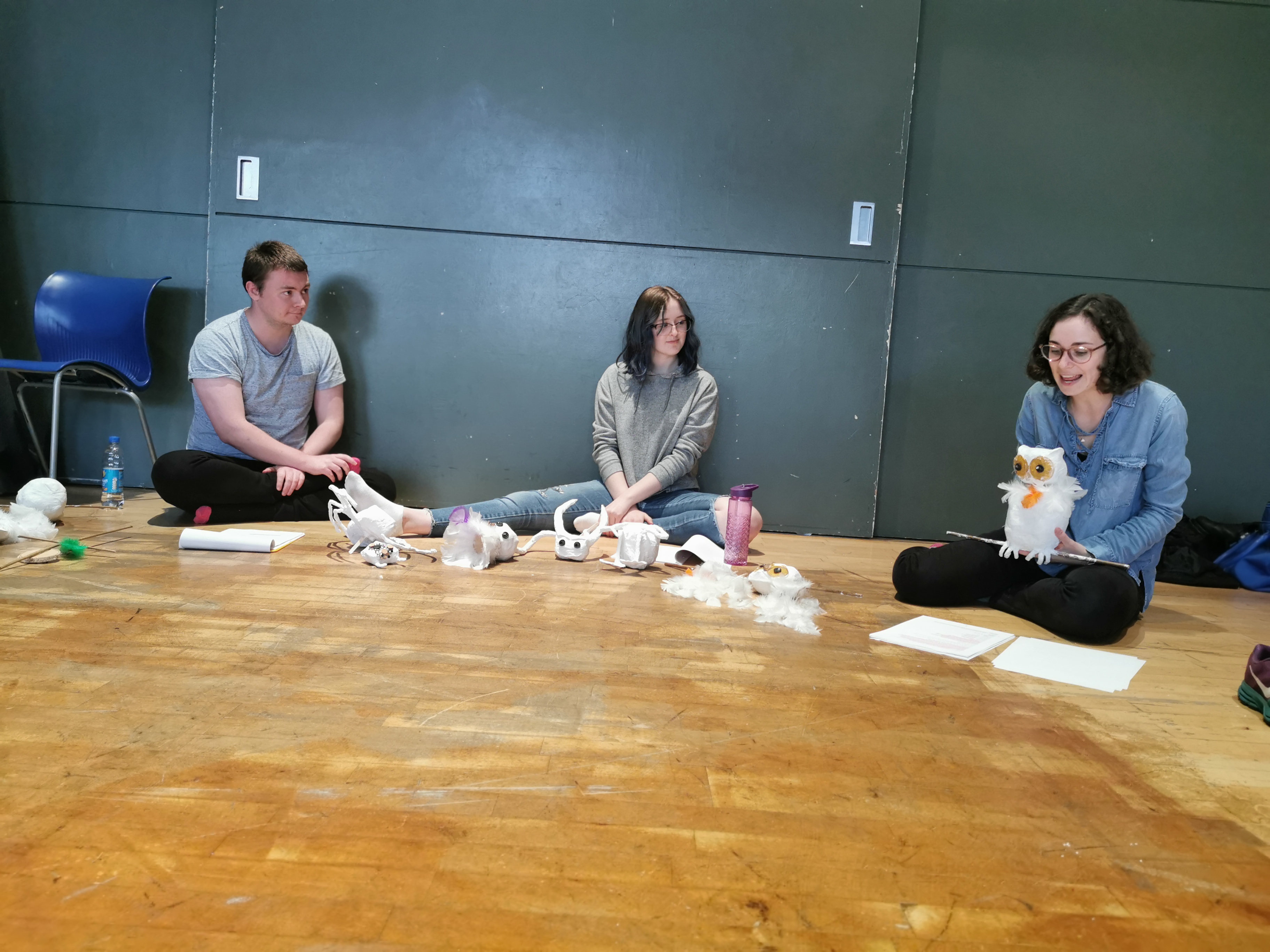 Cast members: John Tulloch, Heather MacDonald and Alina Ben Larbi sit in a semi-circle with scripts and puppet parts surrounding them. John and Heather listen to Alina as she narrates in the voice of a scared owl. The Owl puppet sits on her lap.