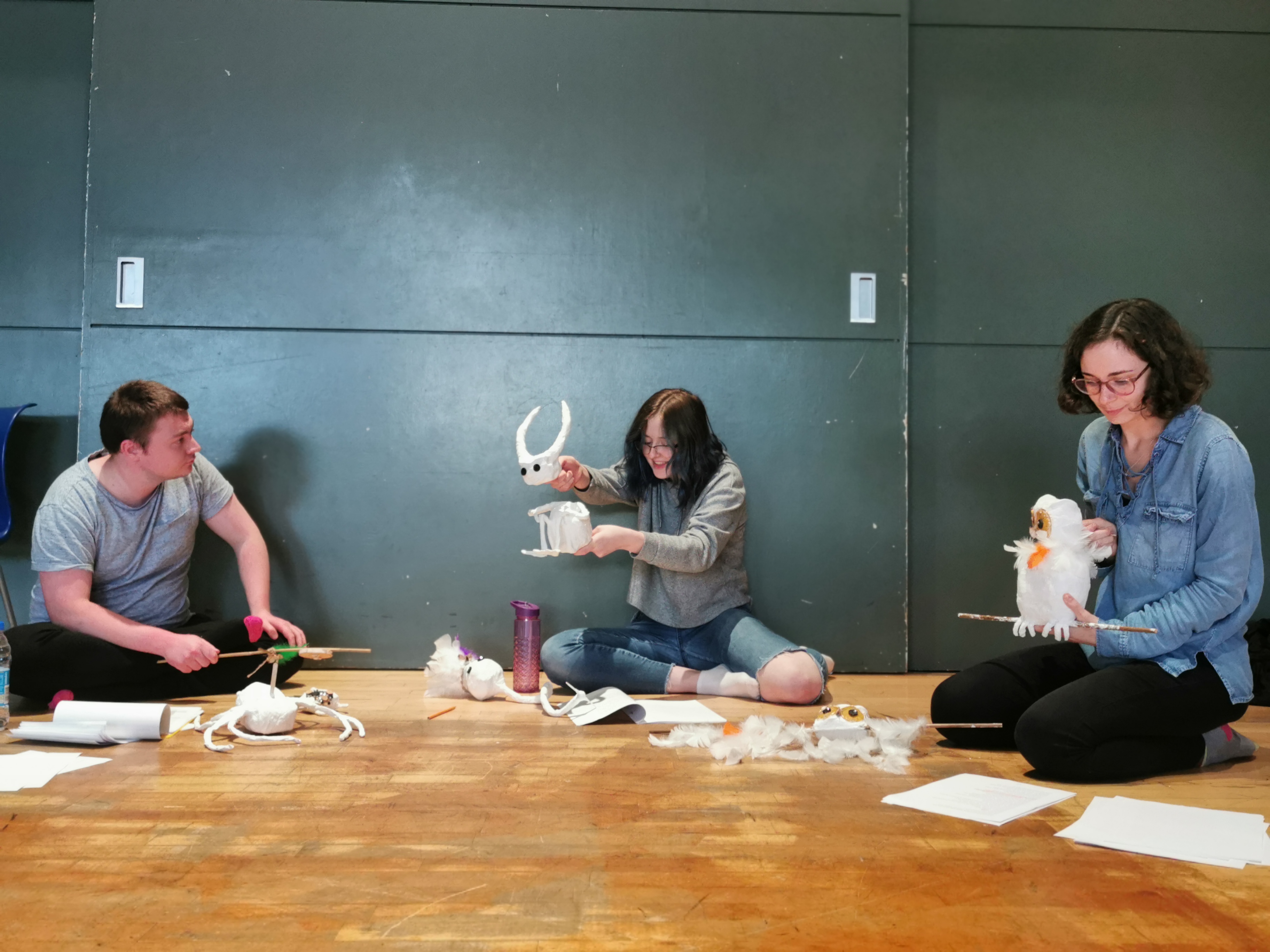 Cast members: John Tulloch, Heather MacDonald and Alina Ben Larbi sit in a semi-circle against a grey wall. Scripts and puppet parts surround them. John listens intently to Heather's narration as the 'imp' puppet (a papier-mâché frog body opertated independently of its large horned head with black button eyes). Heather holds the 'owl' puppet, who looks as if also listening to the imp, while she herself follows the script on the floor.