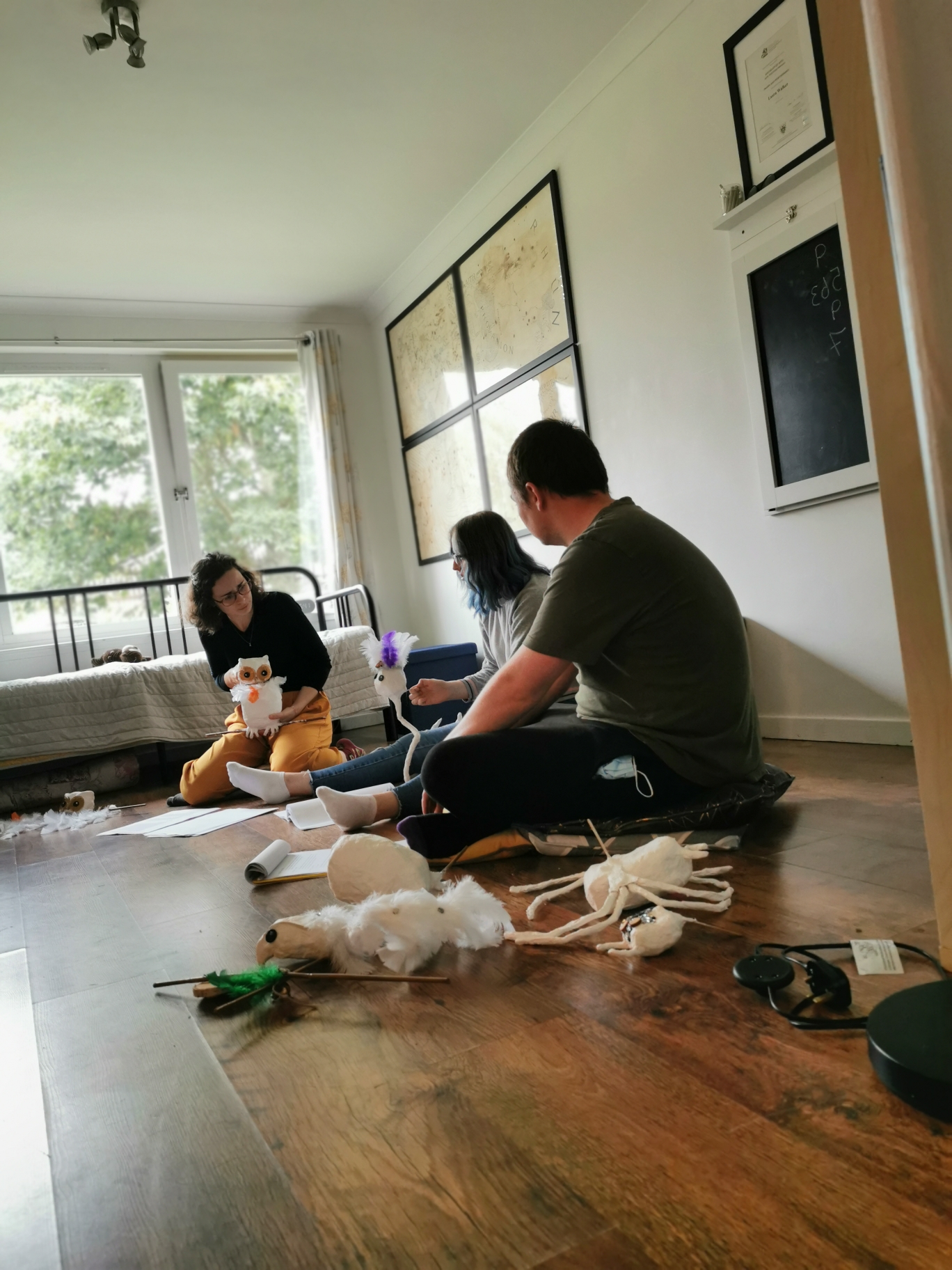 Cast members: John Tulloch, Heather MacDonald and Alina Ben Larbi sit in a semi-circle with scripts and puppet parts surrounding them. Alina, the farthest away, narrates with the owl puppet. Heather, in the middle narrates with the thistle puppet. John, with his back slightly to the camera, listens. At the far end of the room is a large window which lets in a lot of light and through which trees are visible. Picture frames and a chalk board hang on the wall to the right.